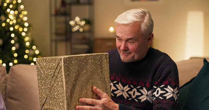 Sitting on the couch, an attractive middle-aged man with gray hair holds a present from his son. Pleasantly surprised and happy, he shook the box and held it to his ear, guessing what was inside. - Powered by Adobe