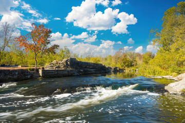 Ros River rushing over rocky rapids into a foamy cascade, framed by vivid autumn foliage and under a bright blue sky dotted with white clouds