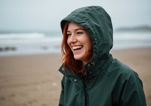 Happy woman with red hair laughs on a beach wearing a green hooded jacket. - Powered by Adobe