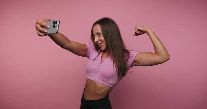 footage of young confident woman flexes her bicep while taking a selfie, showcasing fitness and self-expression on pink background