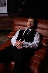 Elegant young man in black suit posing in a modern studio interior with bookshelves and leather sofa, stylish business portrait