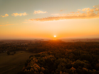 Golden hour aerial view of a forest in autumn, bathed in warm sunlight as the sun sets on the horizon. The vivid orange sky and colorful treetops create a peaceful and cinematic atmosphere, capturing 