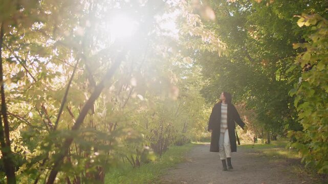 Joyful walker with headphones holds autumn leaf, listens to music and spins beneath sunlit arboretum canopy, relaxed stroll along leafy path, playful turn, serene mood, casual coat boots style