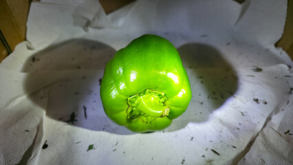 Fresh green bell pepper on white fabric background with scattered dry leaves, minimal food still life, natural texture contrast, simple organic composition.