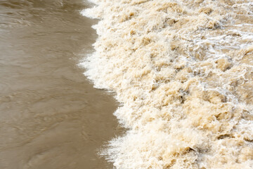 Turbulent muddy flood water with white foam and bubbles creating dramatic texture during extreme weather event. Concept for natural disaster documentation and climate change impact visualization.