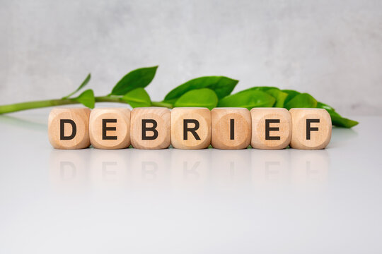 Wooden cubes spelling DEBRIEF on glossy white desk with green leaves in blurred background
