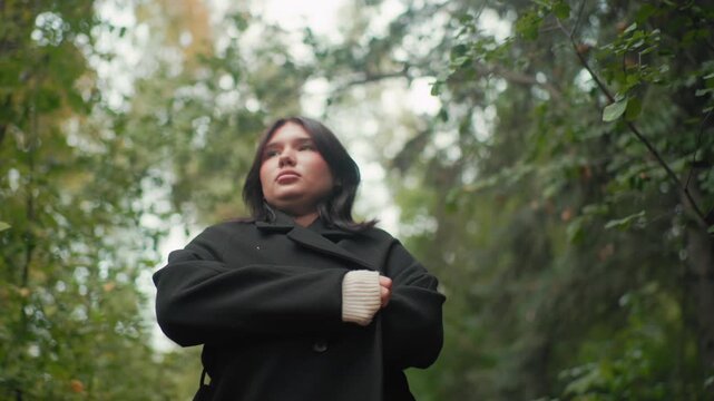 low angle autumn visitor pulling coat close, covering herself with blanket while strolling through forest, chilly day mood, soft bokeh leaves above, calm wander among trees, cozy movement