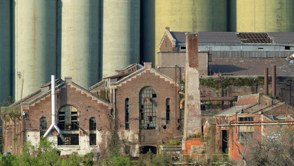 Dilapidated brick industrial buildings with large silos in the background. A scene of urban decay and forgotten industry, overgrown with vegetation