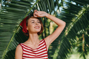 A cheerful woman wearing a striped red and white top poses among lush palm leaves, smiling warmly. Her sunny mood creates a bright, tropical atmosphere and carefree summer vibe.