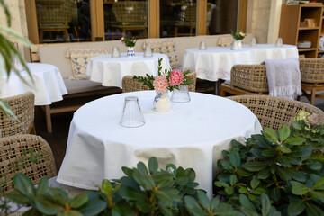 A cozy lounge area in a cafe on an open veranda with chairs and tables. Lush green plants surround the space, creating a relaxing atmosphere.