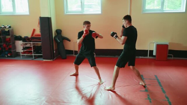 Two wrestlers practicing sparring in gym on red mat, standing barefoot in combat stance, wearing gloves, focusing on martial arts training, building endurance, fighting technique under natural light