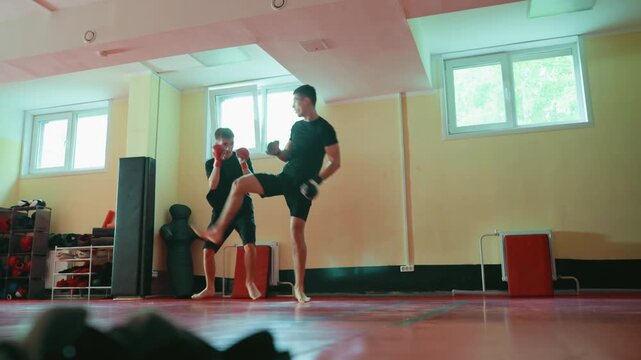 Two fighters sparring in gym on red mat, barefoot, wearing gloves, practicing martial arts techniques, extending punches with focus on combat training