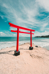 A bright red Torii gate stands prominently on a sandy, rocky outcrop overlooking the turquoise ocean and a distant beach under a vibrant sky.