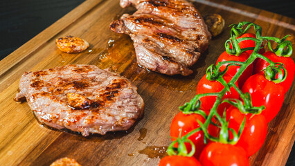 A whole, juicy grilled steak rests on a wooden board, accompanied by roasted cherry tomatoes on the vine and whole fried garlic cloves. Rustic and appetizing culinary scene.