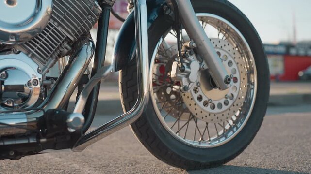 Lower angle close up of motorcycle front wheel and brake disc with rider placing leg on bike stand, chrome engine and metal parts reflecting light, urban street background