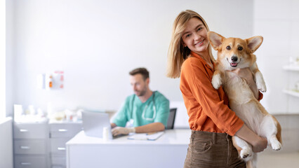 Positive woman owner holding her Pembroke Welsh Corgi dog, being satisfied receiving care from male veterinarian in clinic, panorama with free space