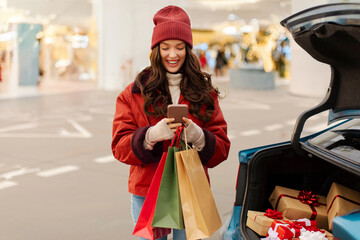 Christmas shopping concept. Happy lady using cellphone and shopper bags standing near open car trunk full of presents outdoors
