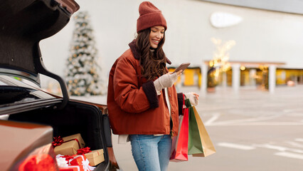 Happy lady checking list of Xmas gifts on cellphone, standing near car with open trunk full of wrapped Christmas presents, outdoor parking © Home-stock