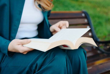 young woman holds thick book in hands, reading book, sitting on bench in park, close-up view, no...