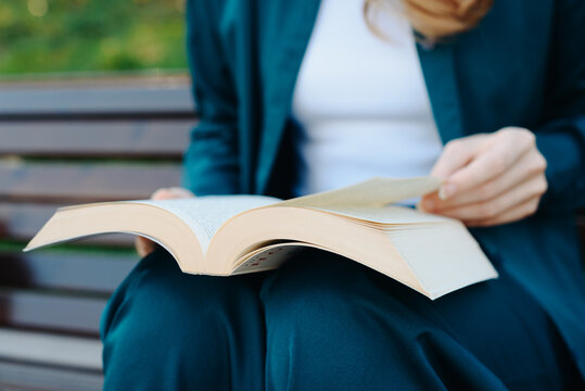 young woman holds thick book in hands, reading book, sitting on bench in park, close-up view, no face