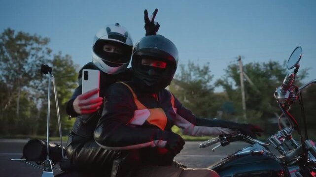 Lady in white helmet takes selfie with husband on motorcycle at sunset, husband in black helmet poses while she makes peace sign gesture, evening road and trees create scenic background
