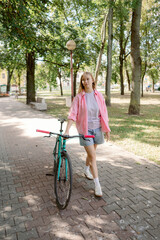 Naklejka premium A girl in a pink jacket and denim shorts strolls down a park path. She pulls her bright bike beside her as she enjoys the sunshine and greenery around her.