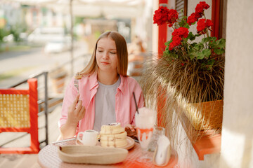 A young woman sits at a small table outside a cafe, enjoying a dessert. She holds a treat in one hand while a plate of pastries and a drink sit nearby among vibrant flowers.