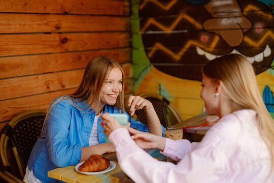 Two young women are sharing a light breakfast at a vibrant cafe. One is smiling while holding a cup, and the other enjoys a croissant. The colorful mural adds charm to the scene.