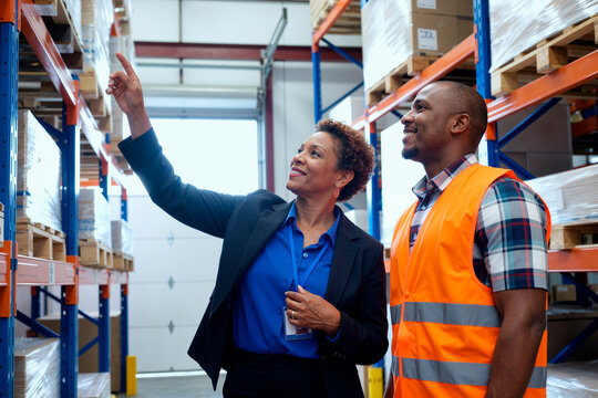 Middle aged Black woman explaining warehouse logistics to young adult Black man wearing safety vest, both standing in industrial storage area surrounded by shelves and pallets