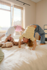Two young girls are performing playful acrobatics on a bed in a bright and cheerful room. They seem to be enjoying their time, laughing and having fun together.