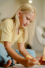 A young girl with long blonde hair sits on the floor, focused on drawing with crayons. She wears a bright yellow top and smiles softly while creating her artwork in a cozy indoor space.