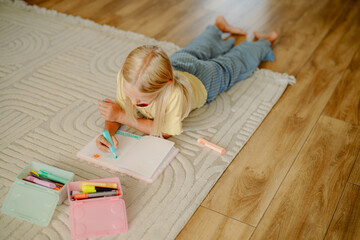 A young girl lies on the floor, using colorful markers to write in her notebook. The cozy living room has warm wooden flooring and a soft area rug.