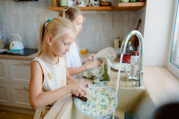 Two young girls are engaged in washing dishes in a bright kitchen. The light streams in through the window, creating a cheerful atmosphere as they clean and dry the dishes together.