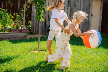 Two children enjoy a bright afternoon in a backyard, playing with a beach ball. One child pushes the other as they run on soft green grass surrounded by blooming flowers.