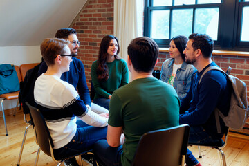 Group of young adult Caucasian and Hispanic men and women sitting in circle engaging in conversation, facing each other in bright room with brick wall and large window