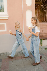 Two young girls walk hand in hand along a charming street, each holding a cone of ice cream. They wear stylish overalls and smile happily while enjoying their treats.