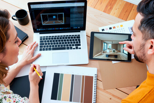 Caucasian young adult woman and Caucasian young adult man collaborating on interior design project, reviewing digital room renderings on laptop and tablet, discussing color palette samples