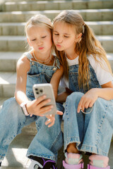 Fototapeta premium Two girls are sitting on stone steps, wearing denim overalls and making funny faces at their phone while capturing selfies. They are enjoying a joyful moment together.