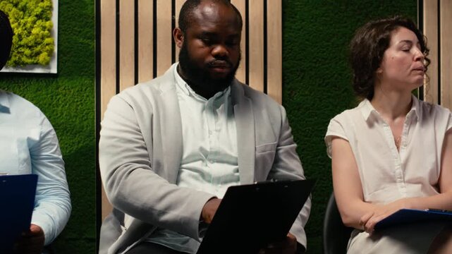 Professional diverse candidates awaiting start of job interview, showcasing anticipation in office hallway, preparing with information for the recruitment process. Career opportunity. Camera B.
