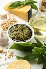 Fresh pesto sauce in bowl and ingredients on white wooden table, closeup