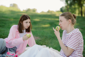 Fototapeta premium In a sunny park, two friends sit on a blanket, savoring fresh apples. One looks at the other thoughtfully, while the other bites into her apple. They relax in the green surroundings.