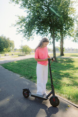 A woman in a pink shirt and white pants stands on an electric scooter in a green park. Bright sunshine illuminates the area, creating a vibrant outdoor atmosphere.