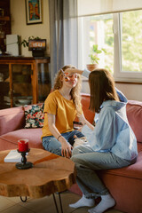 Obraz premium In a bright living room, two friends relax on a soft couch. One wears sunglasses and smiles while the other playfully adjusts her hair. Sunshine fills the room, creating a warm atmosphere.