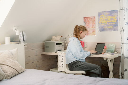 A student sits at a desk in a bright, cozy bedroom, focused on a laptop. The space features a printer, colorful posters, and natural light streaming through the window.