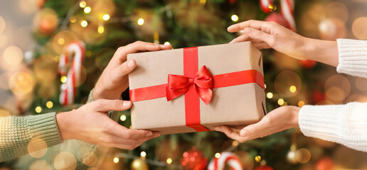 Hands of young man and woman with gift box near Christmas tree at home, closeup