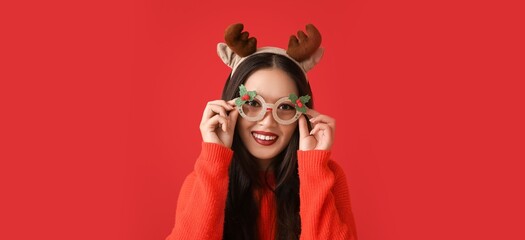 Young Asian woman wearing Christmas glasses and reindeer antlers on red background