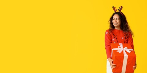 Happy young African-American woman holding big Christmas gift box on yellow background with space for text