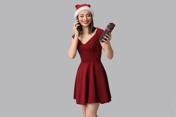 Young woman in Santa hat talking by mobile phone and holding Christmas gift box on grey background