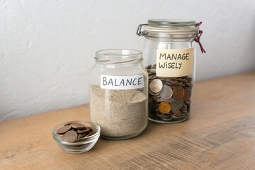 Jars labeled 'Balance' and 'Manage Wisely' filled with coins and sand on a wooden surface
