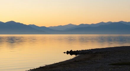 Serene mountain range reflected in a calm lake at sunrise with a rocky shoreline in the foreground.